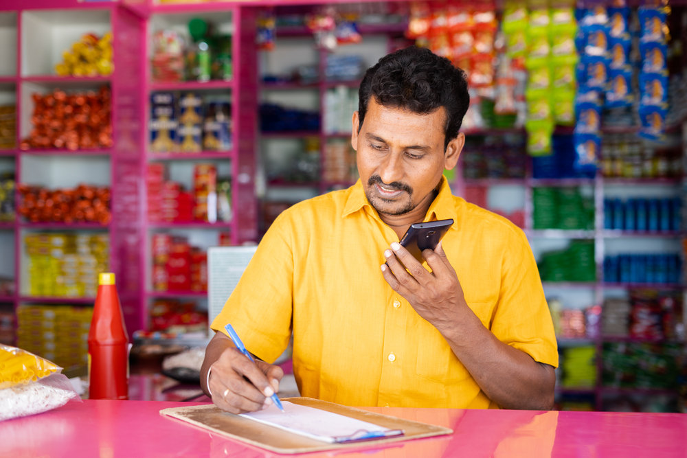 Indian groceries business man noting orders while talking with customer on mobile phone at kirana shop - concept of distance shopping - home delivery service borzo